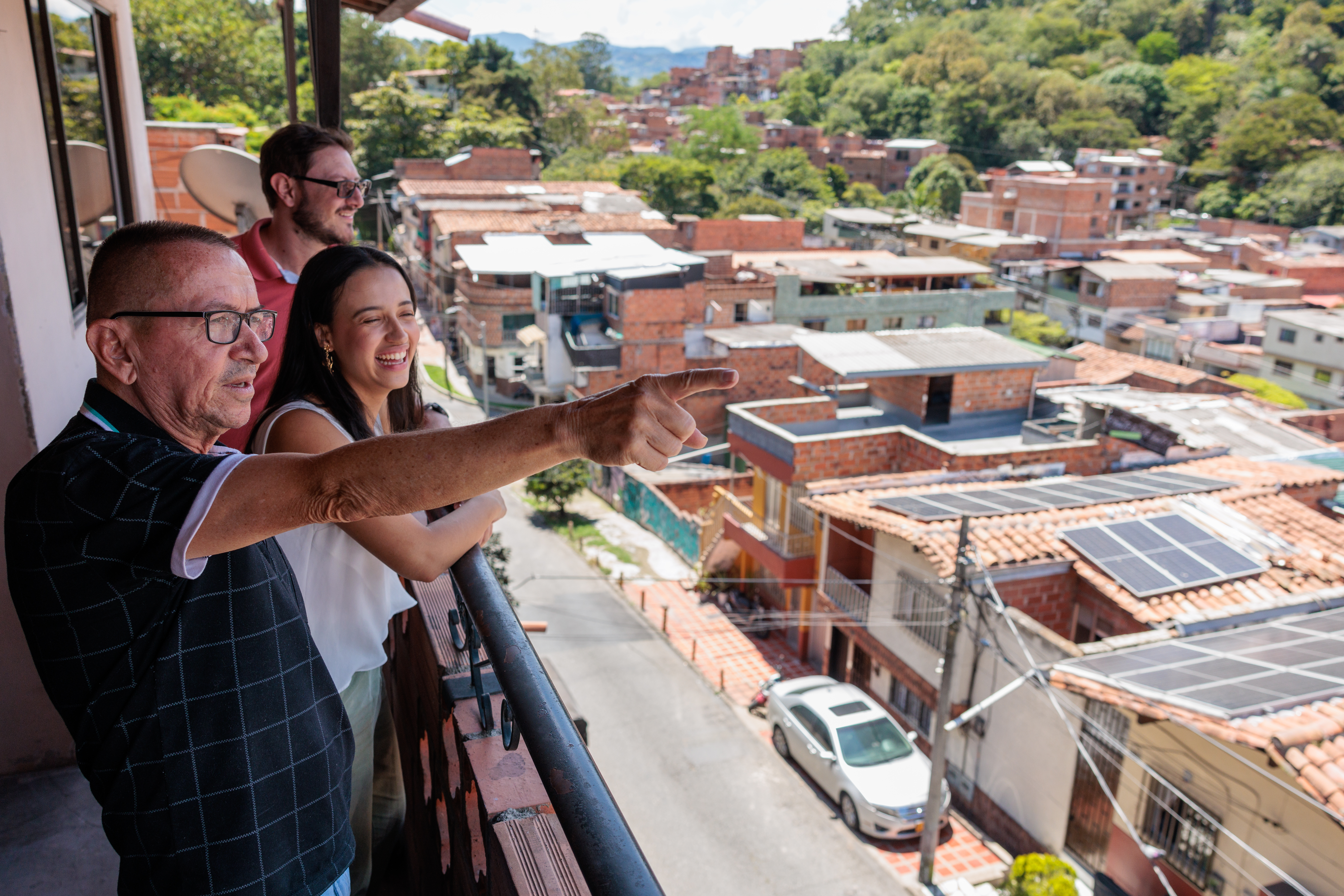 A man on a balcony points out over a residential area of homes with solar panels on their roofs, speaking to a woman and man to his left, both smiling.