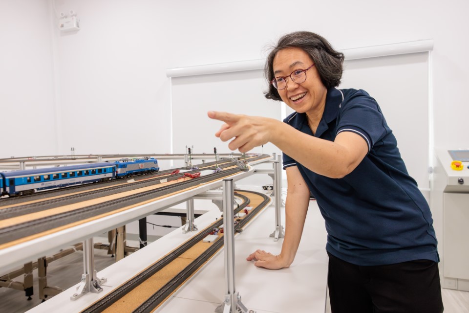 Smiling Woman in laboratory setting points to a blue train on a public transport model