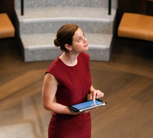 A woman in a sleeveless red dress looks up to towards to top right. In her hands she holds an ipad.