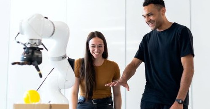 A man gestures towards a robotic arm. A woman beside him is smiling and laughing. 